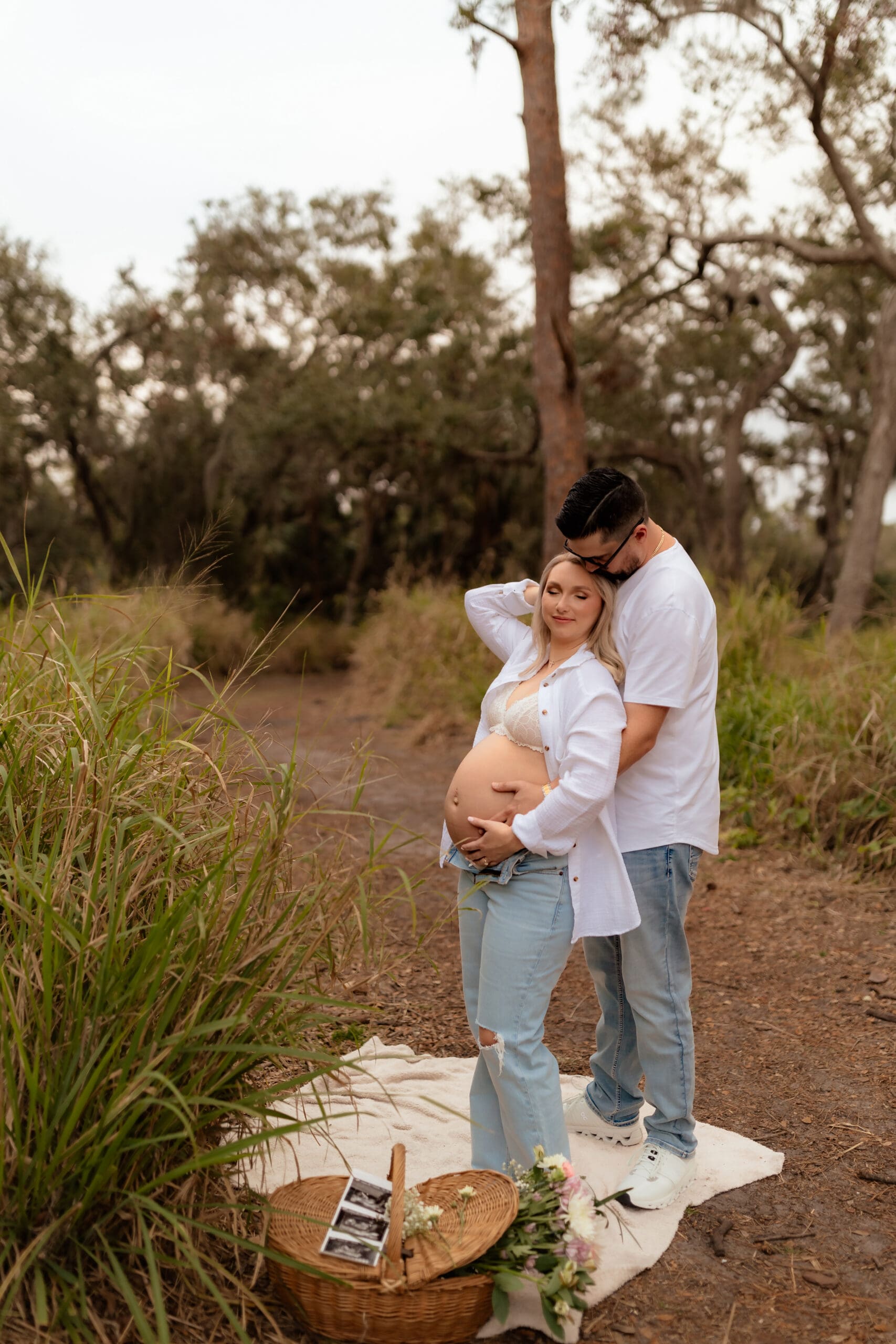 Couple Holding Baby Bump with Wildflowers – A warm, intimate outdoor maternity photo of a couple embracing, with the father-to-be gently holding the baby bump while small wildflowers peek from the mother’s jeans pocket.